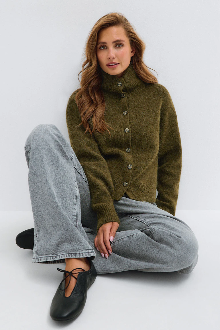 Olivgrüne Strickjacke mit hohem Kragen und Knopfleiste, hellgraue Jeans und schwarze Schnürballerinas.