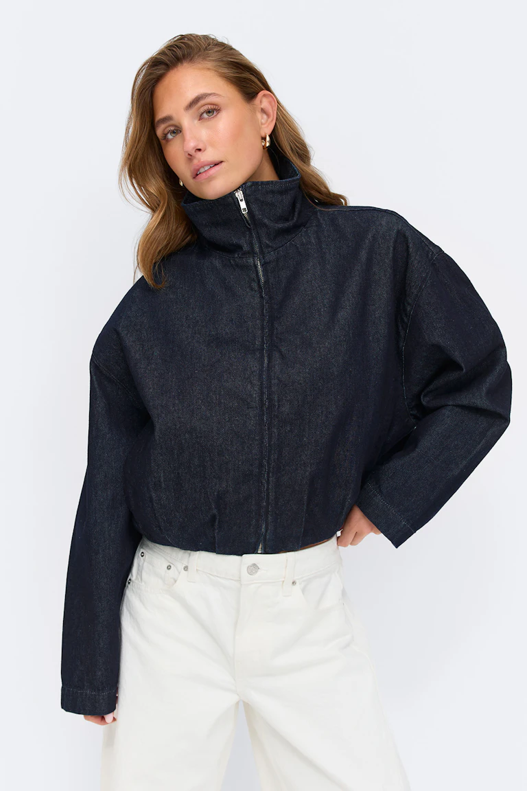 Dunkle indigoblaue Jeansjacke mit Reißverschluss, hohem Trichterkragen und langen Ärmeln, kombiniert mit weißen Jeans mit geradem Bein und hoher Taille.
