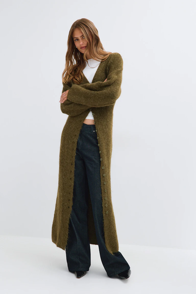 Long olive-green knit cardigan over a white top, paired with dark wide-leg jeans and black shoes.