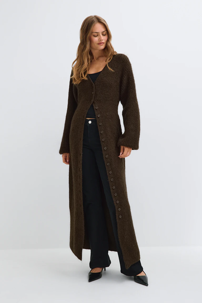 Un long cardigan en maille marron foncé porté par-dessus un haut noir, associé à un pantalon noir à jambes larges et à des escarpins noirs à bout pointu.
