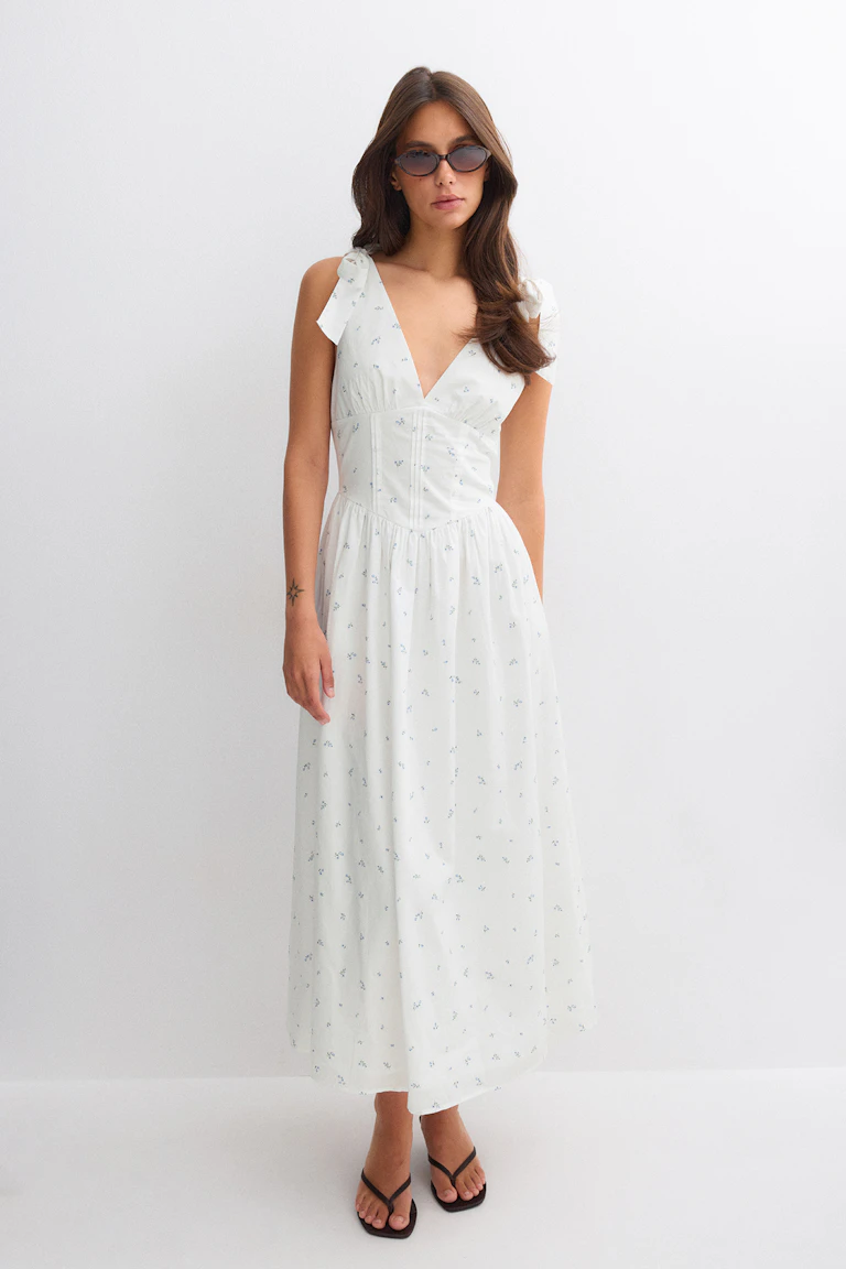 Weißes Maxikleid mit kleinem blauen Blumenmuster, tiefem V-Ausschnitt, geschnürten Schulterträgern und tailliertem Oberteil, kombiniert mit einer schwarzen Sonnenbrille und schwarzen Zehenstegsandalen.