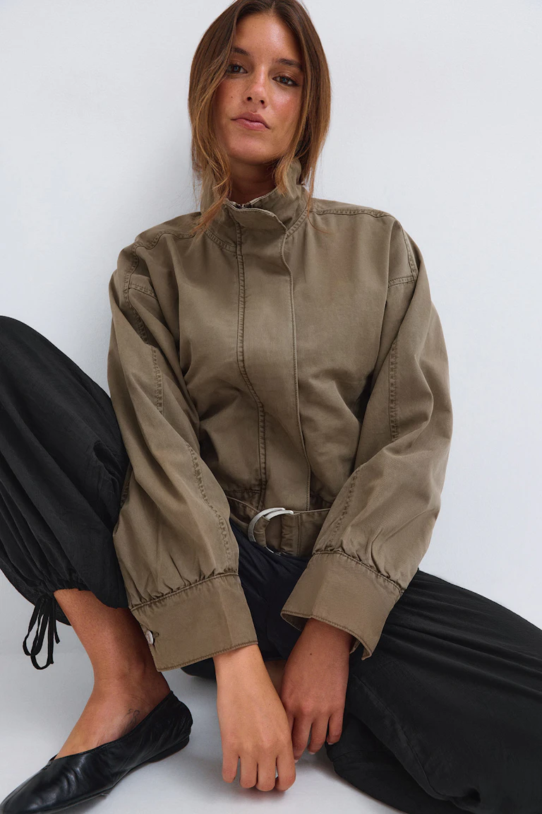 Eine Frau, die eine khakifarbene Jacke mit hohem Kragen und Gürtel, eine schwarze Plisseehose und schwarze Ballerinas trägt.