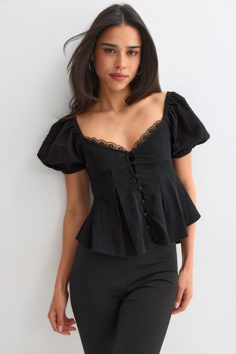 Black textured peplum top with puff short sleeves, lace‑trimmed sweetheart neckline and fabric‑covered front buttons, paired with black fitted trousers.