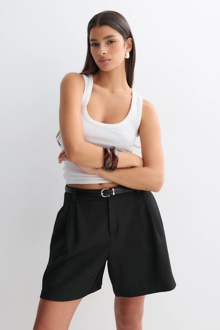 White ribbed scoop-neck tank top paired with high-waisted black tailored Bermuda shorts and a slim black belt with silver buckle, accessorized with tortoiseshell bangles and white textured hoop earrings.