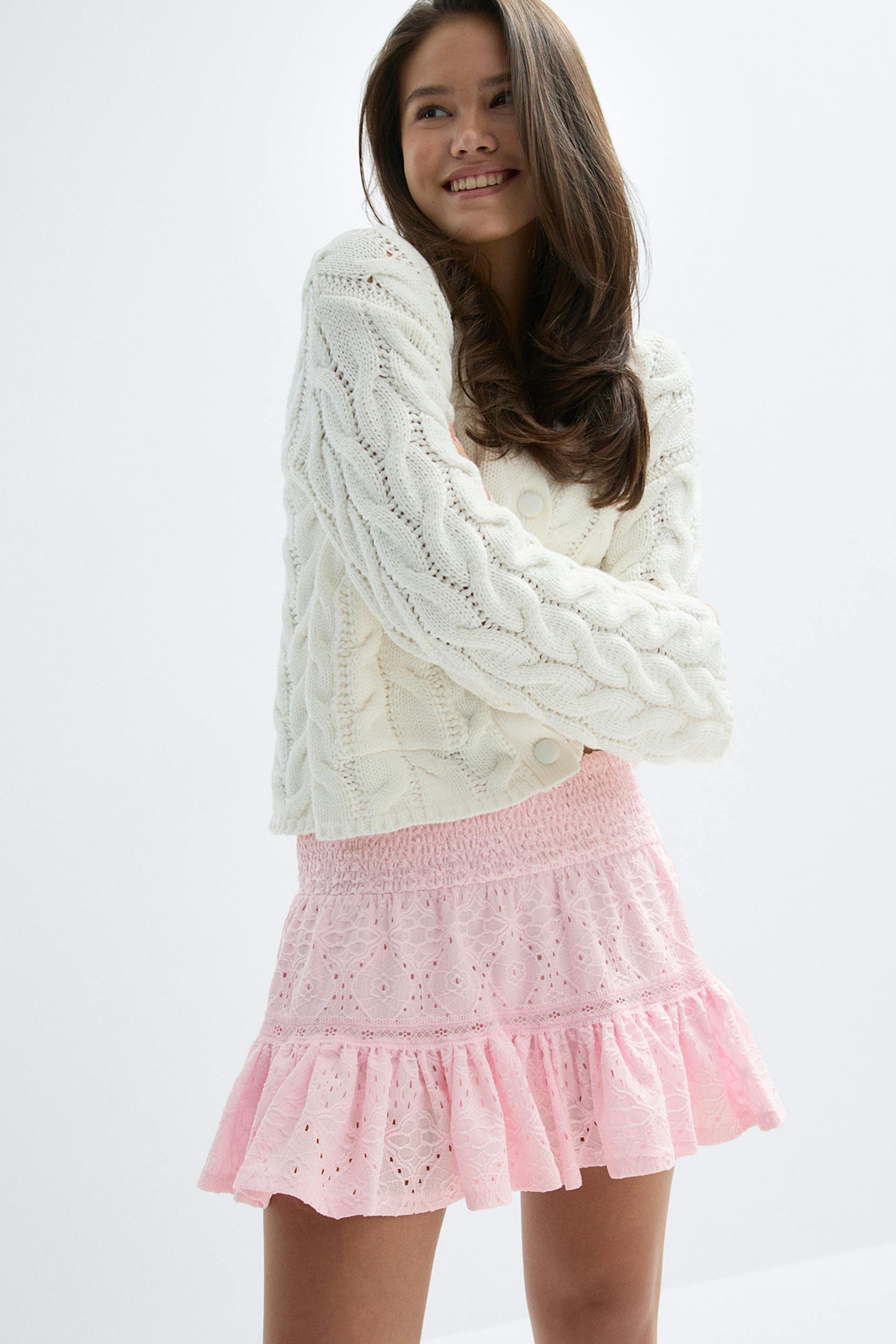 Cardigan boutonné en tricot câblé crème à manches longues, associé à une mini-jupe rose en dentelle œillets avec taille smockée et ourlet volanté.