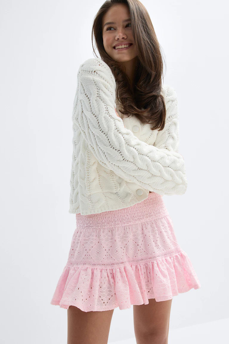 Cardigan boutonné en tricot câblé crème à manches longues, associé à une mini-jupe rose en dentelle œillets avec taille smockée et ourlet volanté.