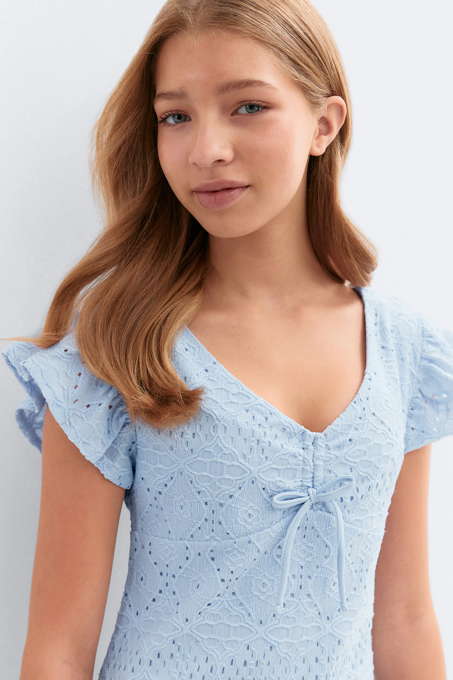 Robe en dentelle à œillets bleu clair, décolleté en V, manches courtes volantées et petit nœud papillon sur le devant.