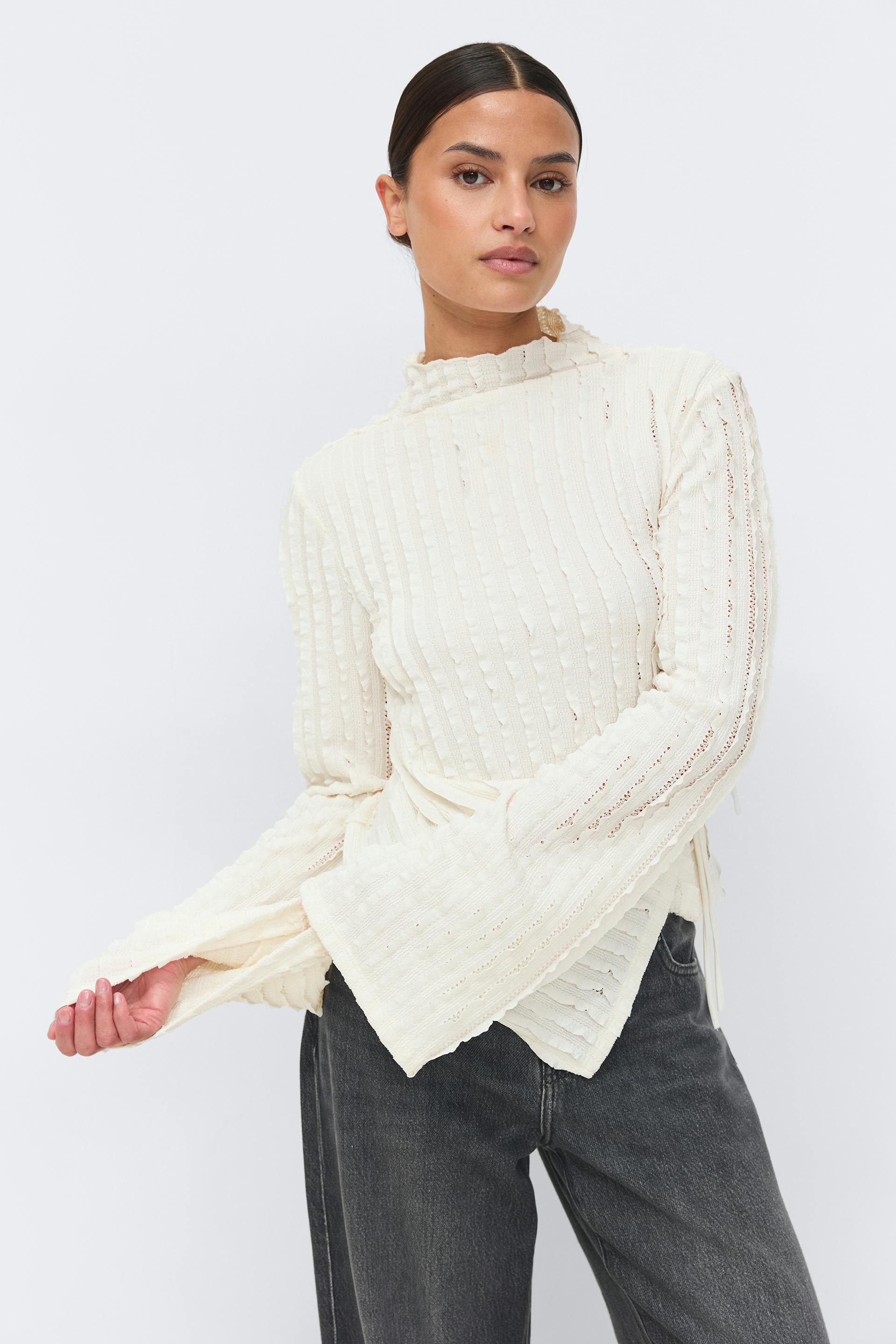 Ivory textured ribbed knit top with high ruffled neckline, openwork vertical stripes, flared bell sleeves and side‑tie wrap hem; paired with high‑waisted charcoal washed jeans.