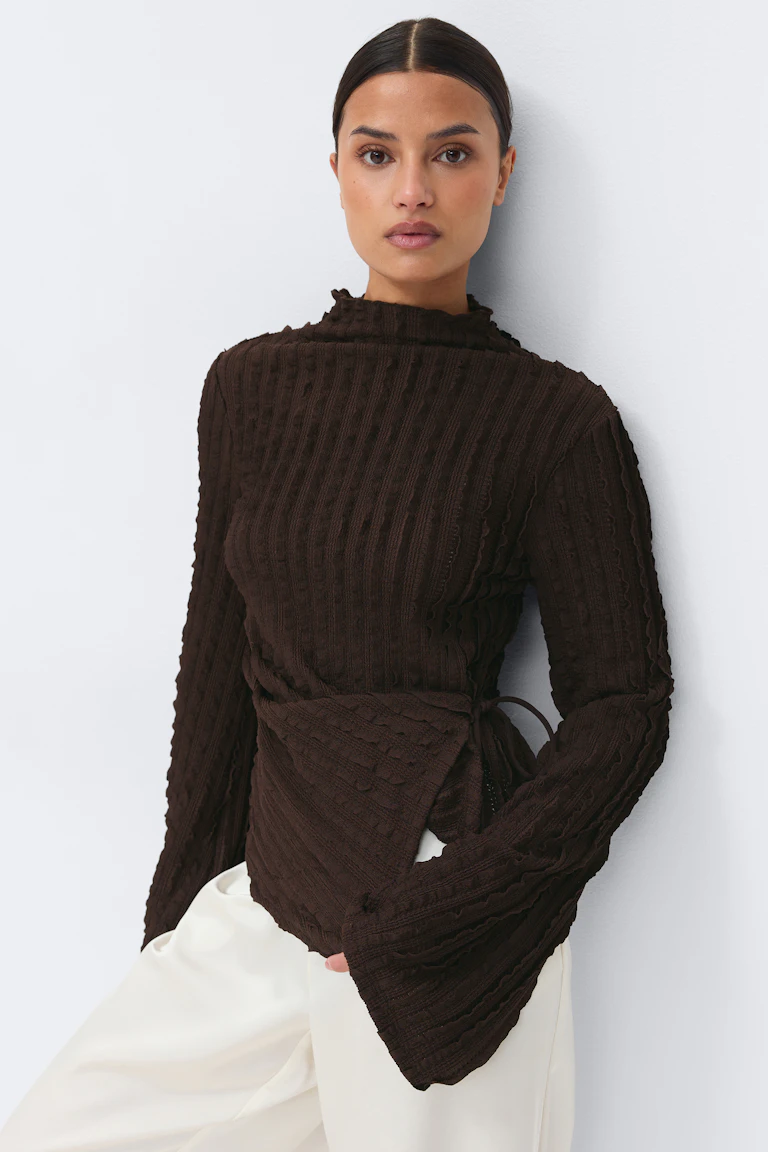 Dunkelbrauner, strukturierter, gerippter Wickelpullover mit langem, ausgestelltem Halsausschnitt, kombiniert mit einer cremefarbenen Hose mit weitem Bein.