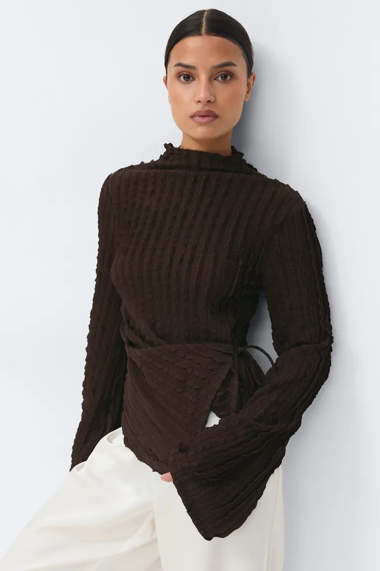 Dunkelbrauner, strukturierter, gerippter Wickelpullover mit langem, ausgestelltem Halsausschnitt, kombiniert mit einer cremefarbenen Hose mit weitem Bein.