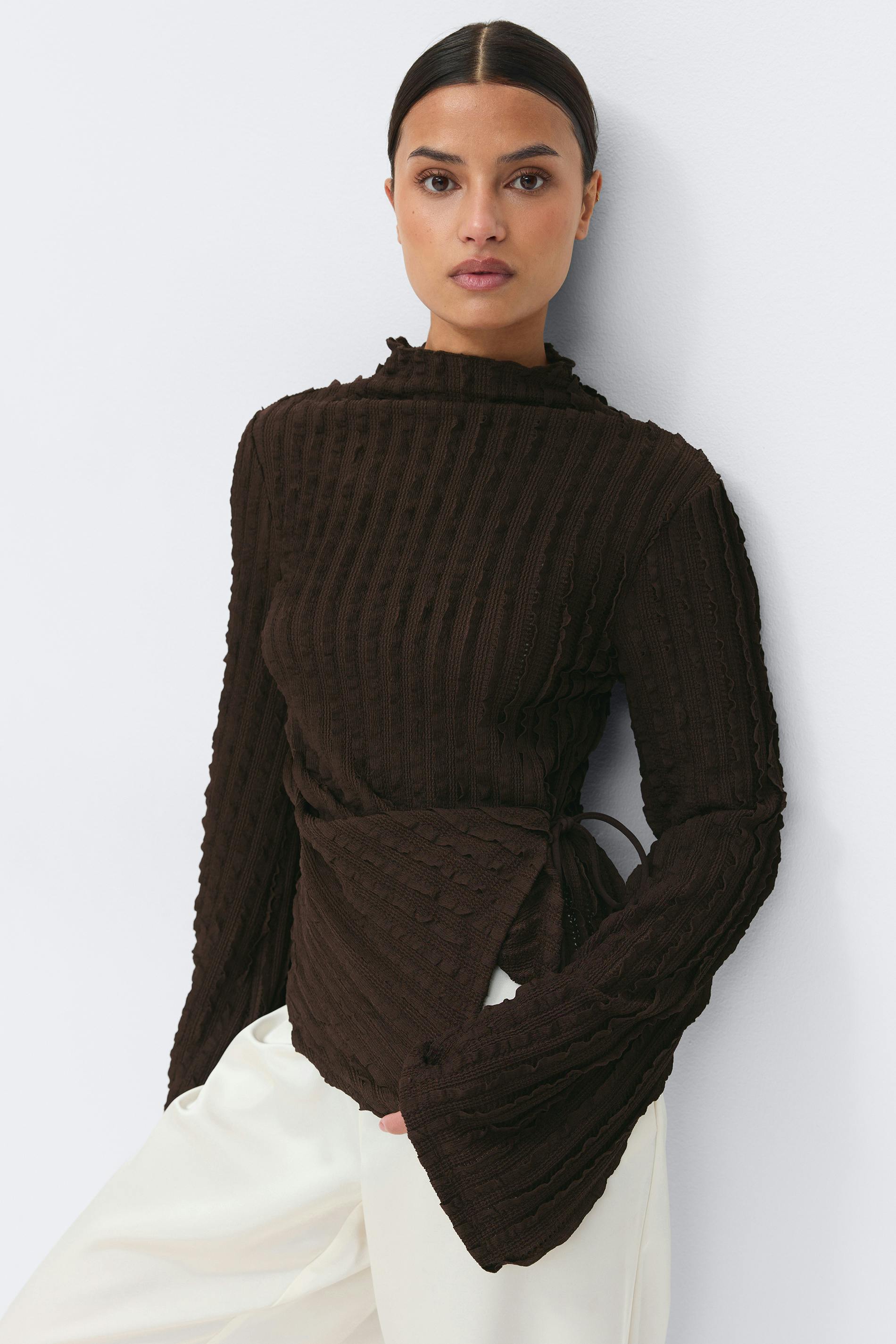 Dark brown textured ribbed mock‑neck wrap sweater with long flared sleeves, paired with off‑white wide‑leg trousers.