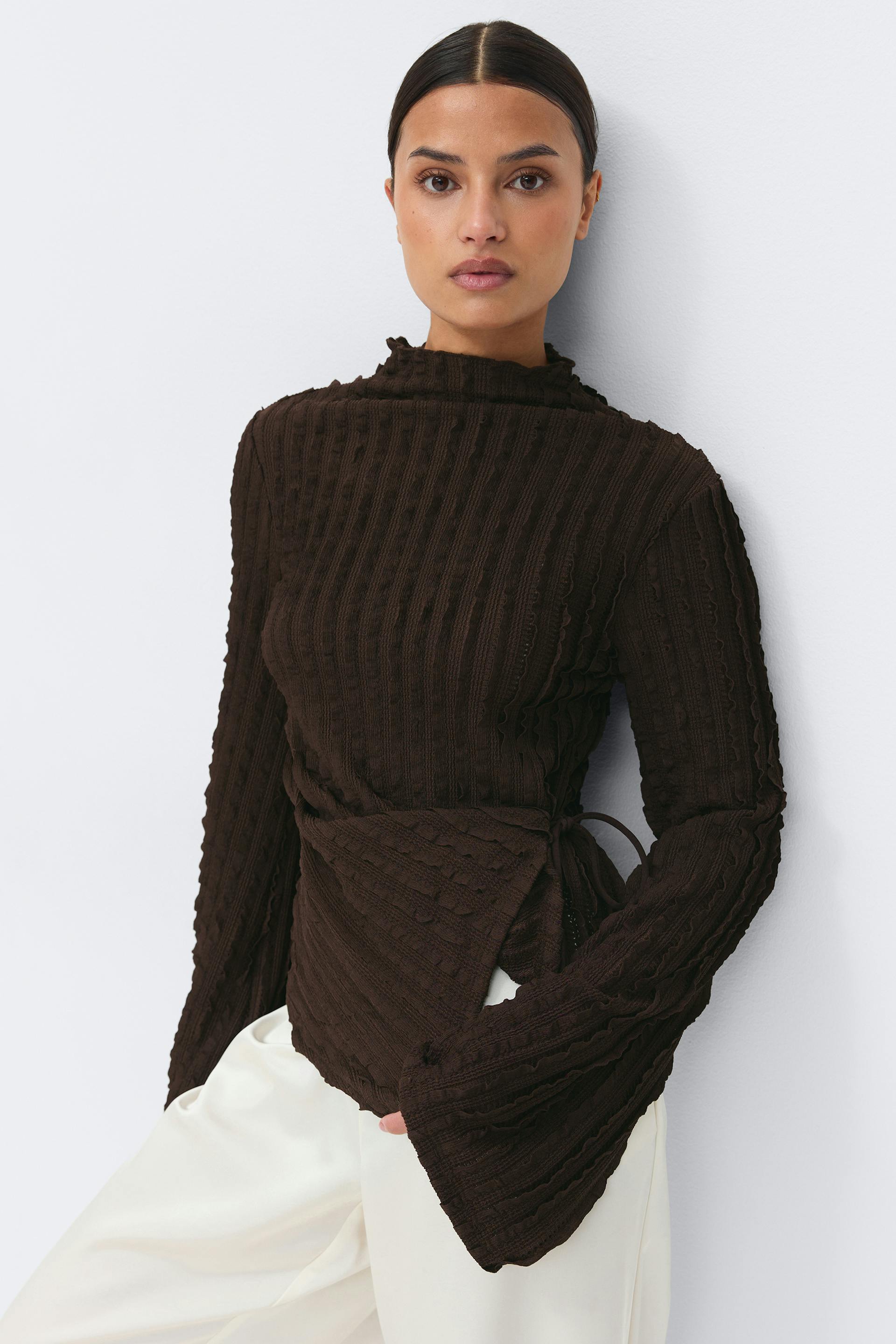 Dark brown textured ribbed mock‑neck wrap sweater with long flared sleeves, paired with off‑white wide‑leg trousers.