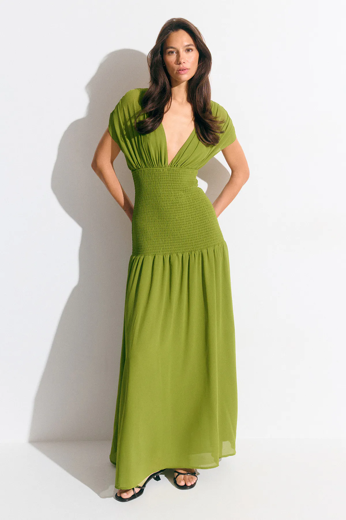 Olivgrünes Maxikleid mit tiefem V-Ausschnitt, kurzen Kappenärmeln, gesmokter hoher Taille und fließendem Rock, kombiniert mit schwarzen Riemchensandalen mit Absatz.