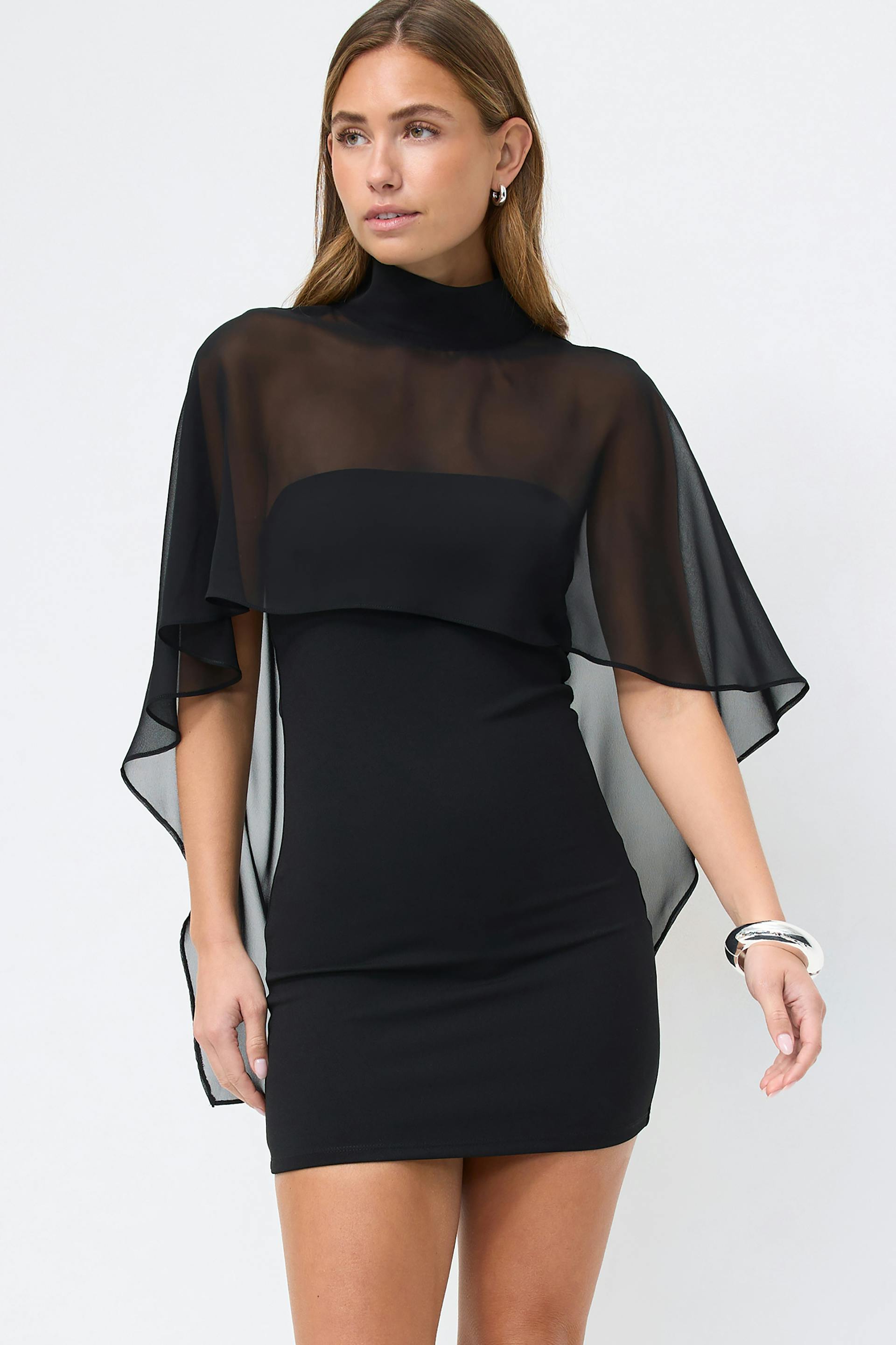 Black fitted mini dress with mock neckline and sheer chiffon cape overlay, paired with a silver cuff bracelet.