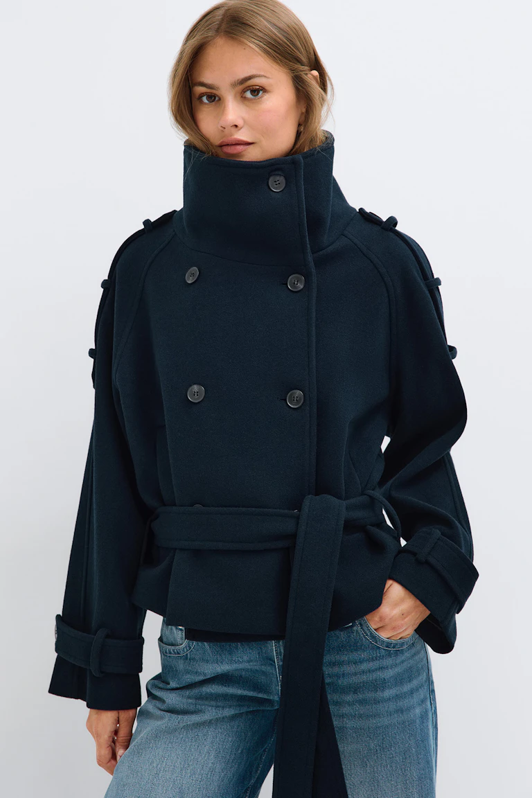 StyleColorSupplier 16657: Marinblå dubbelknäppt kappa med hög krage, bälte i midjan och knäppta manschetter, i kombination med blå jeans.