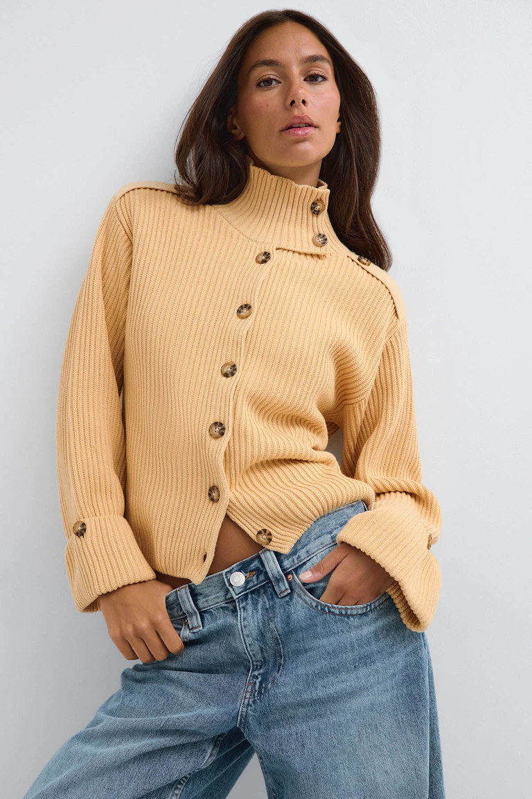 Beige geribbeld gebreid vest met hoge hals en knoopjes, gecombineerd met een blauwe jeans met wijde pijpen.