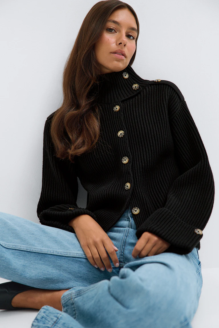 Un pull à col roulé noir côtelé boutonné, associé à un jean bleu clair et des chaussures noires.