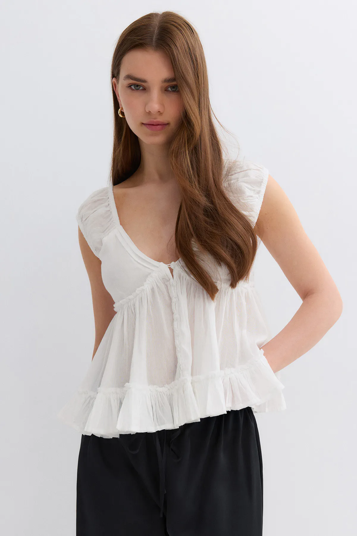 Chemisier blanc à volants sans manches, à encolure en V profond et boutonné sur le devant, associé à un pantalon large noir à cordon de serrage et à de petites boucles d'oreilles dorées.