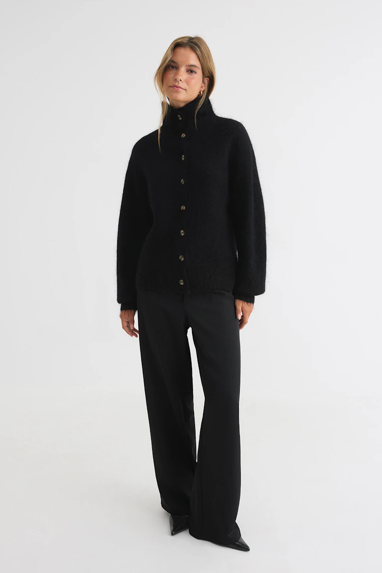 330417 | 17900: Schwarze, hochgeschlossene Strickjacke mit Knöpfen, Hose mit weitem Bein und spitze Schuhe.