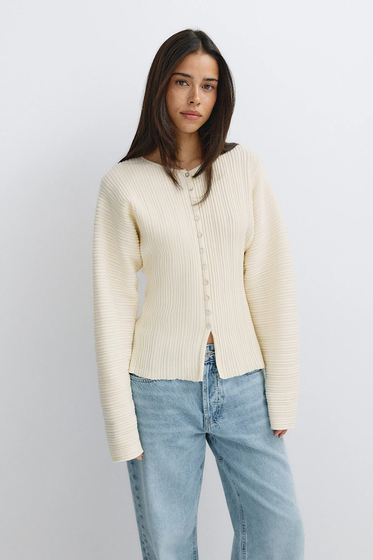 Cremefarbene, gerippte Strickjacke mit Knopfleiste, langen, voluminösen Ärmeln und einem kleinen Schlitz am vorderen Saum, kombiniert mit hell gewaschenen Jeans mit geradem Bein.