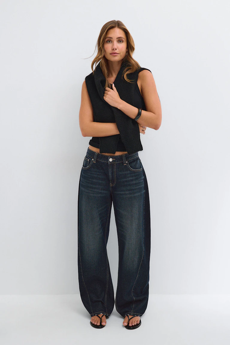 Schwarzes, ärmelloses Oberteil mit einem schwarzen Schal, dunkle Jeans mit weitem Bein und schwarze Sandalen.
