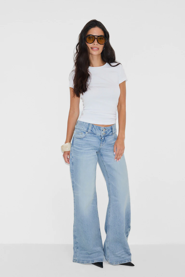 Wit crewneck T-shirt met korte mouwen; lichtgekleurde jeans met hoge taille en wijde pijpen met dubbele knopen; zwarte hakken met spitse teen; oversized bruin getinte zonnebril; dikke ivoorkleurige armband.