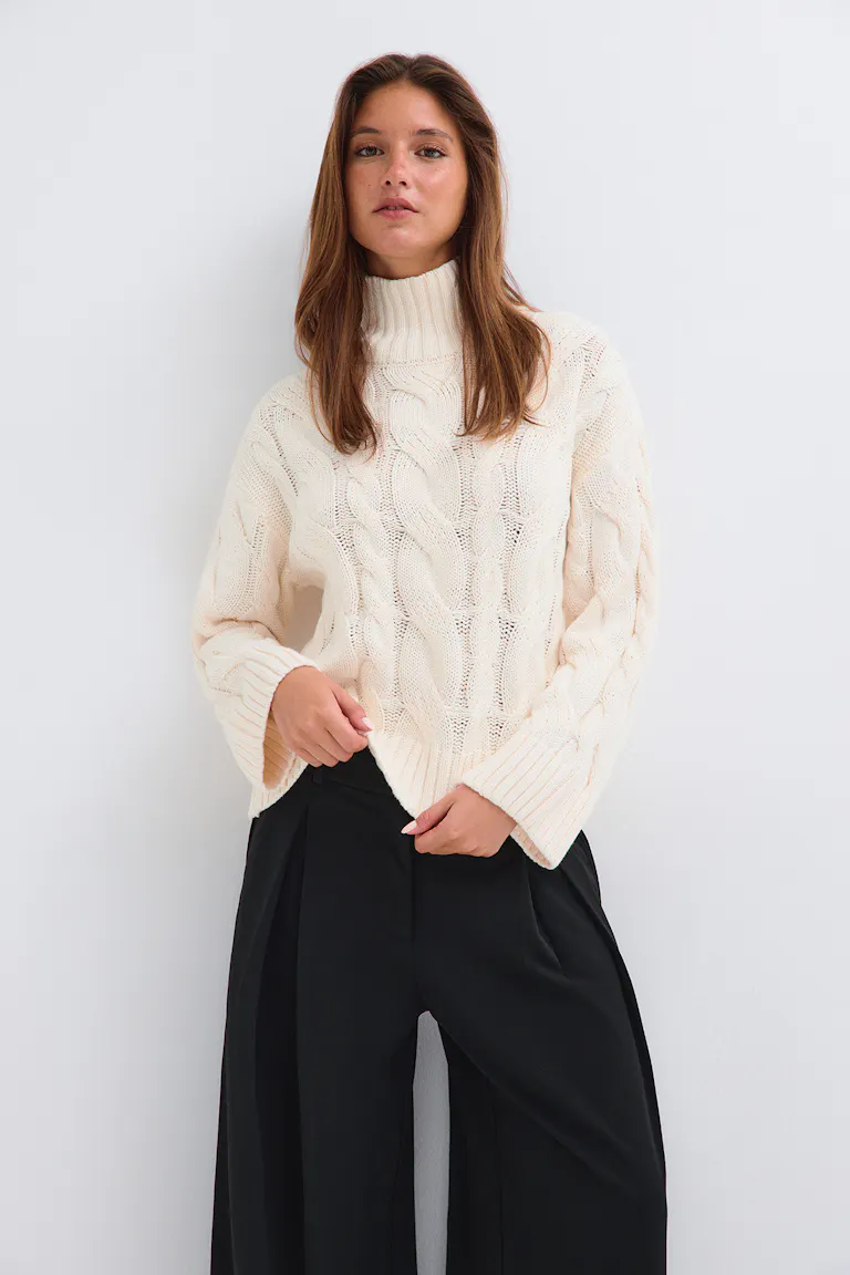 Cremefarbener Rollkragenpullover im Zopfmuster mit schwarzer Hose mit weitem Bein.