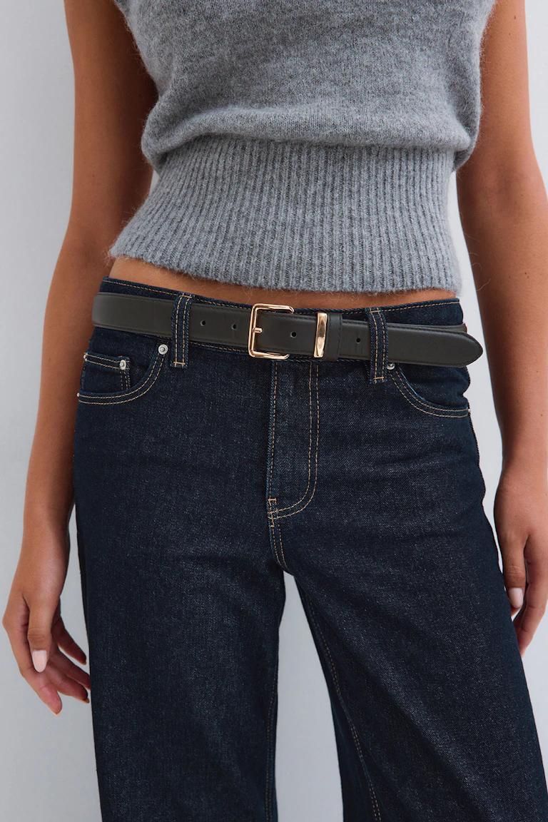 Un haut en maille gris sans manches avec un ourlet côtelé, un jean foncé à coupe droite et une ceinture noire à boucle dorée.