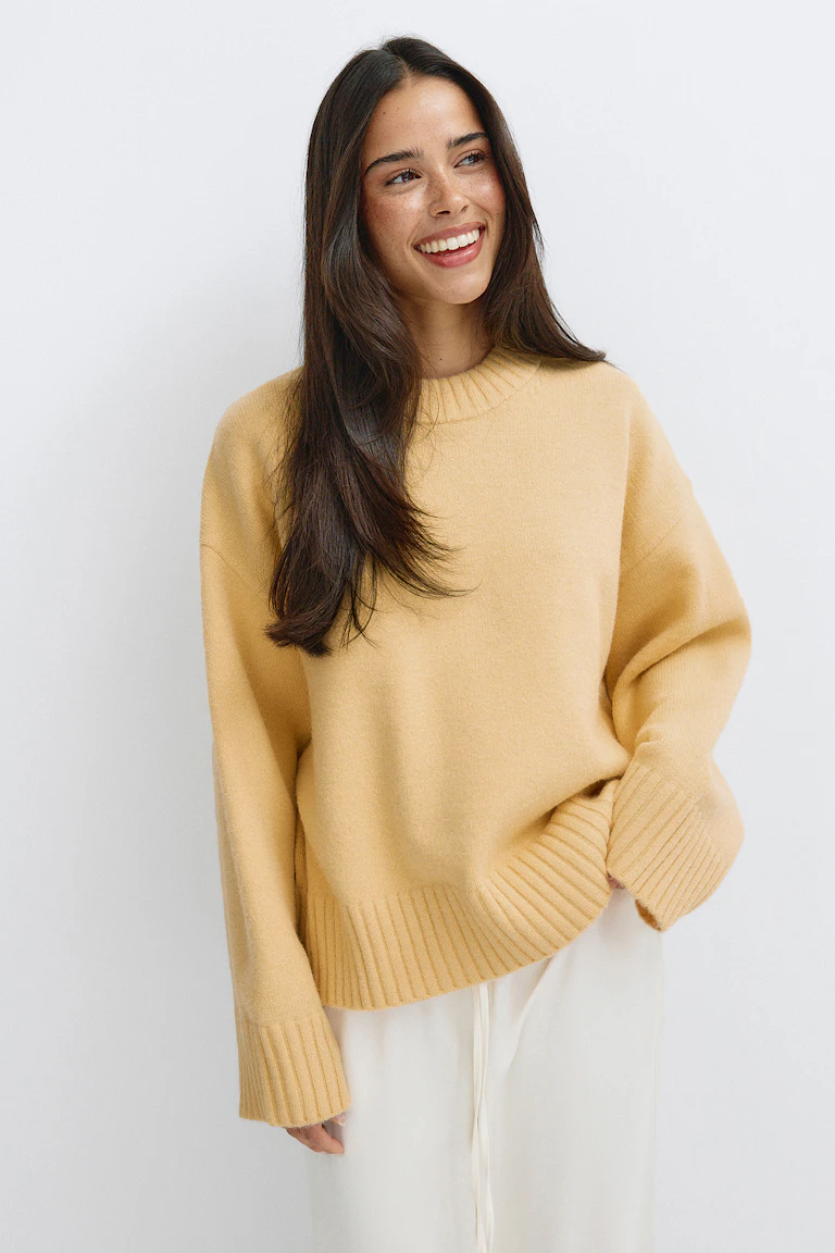 Oversize-Pullover in Pfirsich-Beige mit Rundhalsausschnitt, gerippten Bündchen und Saum, kombiniert mit einer weißen Hose mit weitem Bein und Kordelzug.