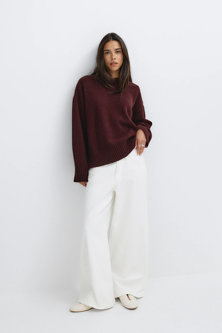 Oversized burgundy crewneck sweater with ribbed hem and cuffs, paired with high-waisted wide-leg white trousers and cream flat shoes.