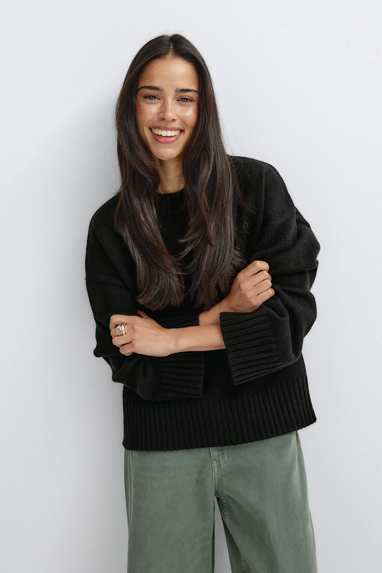 Schwarzer, übergroßer Strickpullover mit gerippten Bündchen und Saum, kombiniert mit einer olivgrünen Hose mit weitem Bein und einem silbernen Ring.