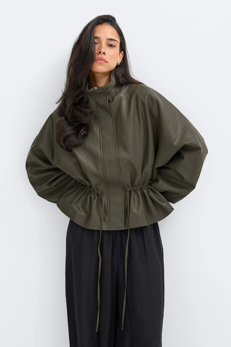 Veste en cuir vert olive à col montant, fermeture éclair dissimulée sur le devant, manches bouffantes volumineuses et cordon de serrage ajustable à la taille, portée avec un pantalon noir ample à jambes larges.
