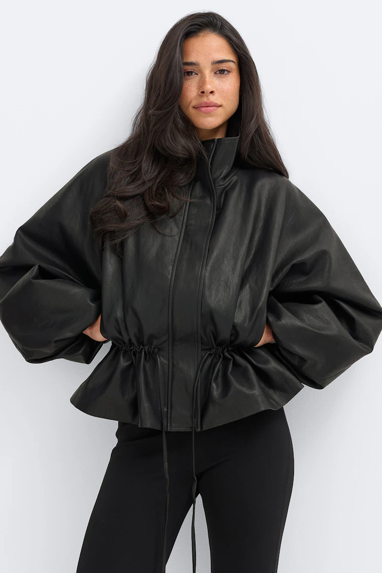 Svart läderjacka med hög trattkrage, överdrivet vida ballongärmar och en midja med peplum som dras åt med dragsko, i kombination med svarta, figursydda leggings.