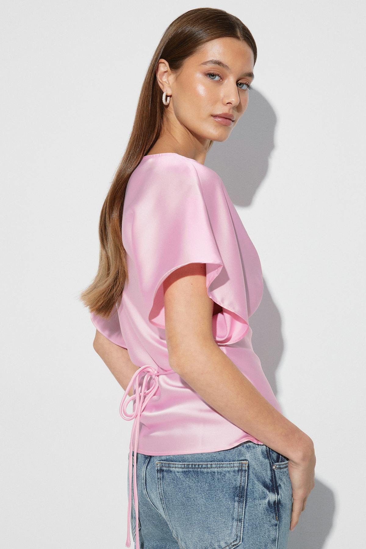 Blouse en satin rose à manches courtes et à taille nouée, associée à un jean clair et à de petites boucles d'oreilles.