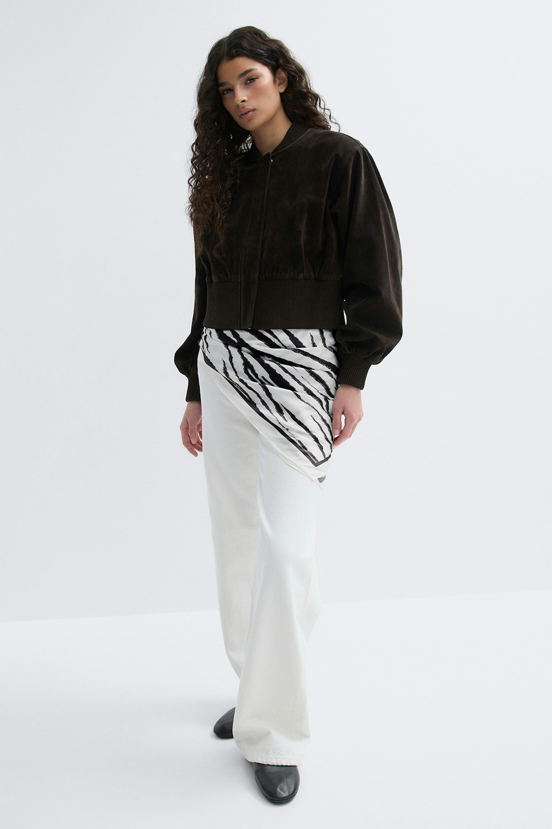 Dunkelbraune Bomberjacke aus Wildleder mit geripptem Kragen, Manschetten und Saum, gepaart mit einem schwarz-weißen Seidenschal mit Zebramuster, der über eine hochtaillierte weiße Hose mit weitem Bein und schwarzen spitzen Schuhen drapiert ist.