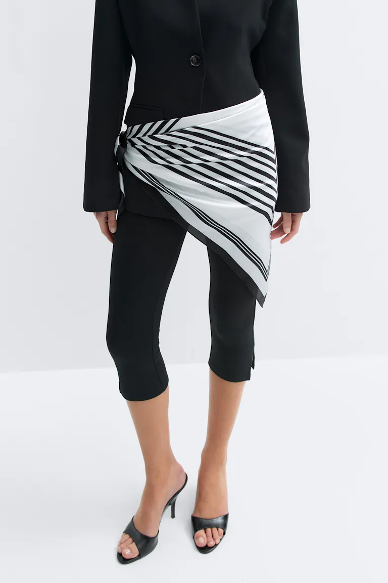 Zwarte getailleerde blazer, zwarte slim-fit broek, zwarte muiltjes met open teen en een witte zijden sjaal met zwarte diagonale strepen die als een asymmetrische sjerp op de heup is geknoopt.
