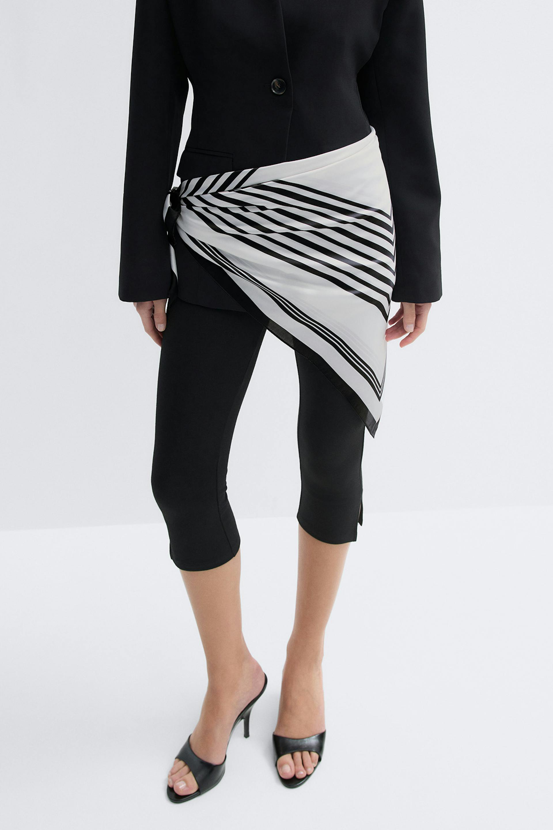 Schwarzer taillierter Blazer, schwarze taillierte Hose, schwarze Pantoletten mit offenen Absätzen und ein weißer Seidenschal mit schwarzen Diagonalstreifen, der an der Hüfte als asymmetrische Schärpe gebunden wird.