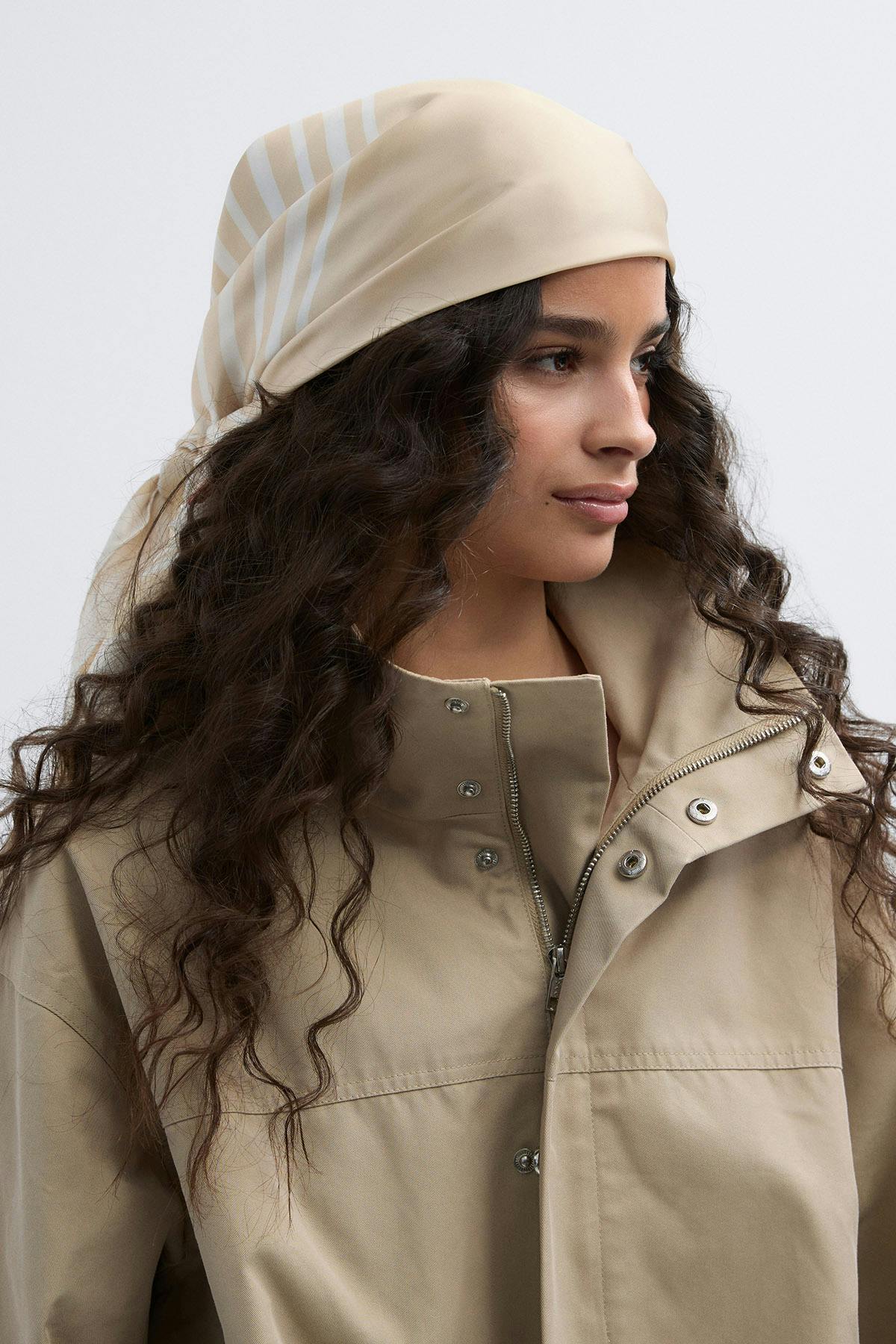 Beigefarbenes Seidenkopftuch mit weißen Streifen, gepaart mit einer beigen Jacke mit hohem Kragen, die mit einem Frontreißverschluss und Druckknöpfen versehen ist.