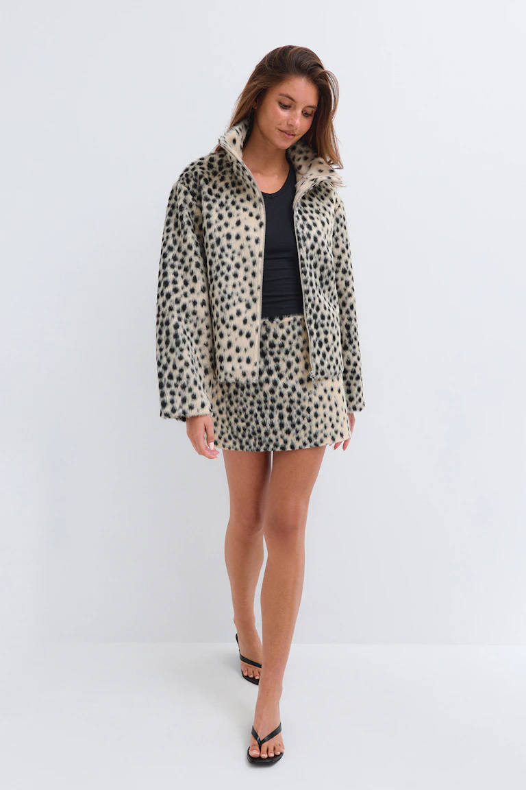 She is wearing a spotted faux-fur jacket and matching mini skirt over a black top, paired with black flip-flop sandals.