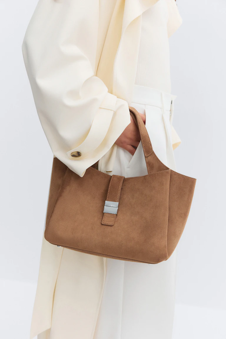 Crèmekleurige jas met lange mouwen over een witte geribde top, gecombineerd met een witte broek met hoge taille en een tan suède tas met korte hengsels en een zilveren gesp.