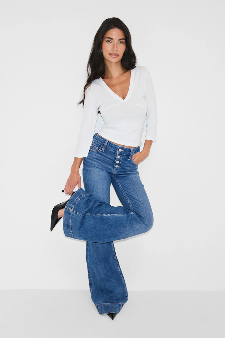 White V-neck fitted top with three-quarter sleeves, paired with high-waisted blue button-fly flared jeans and black pointed-toe heels.
