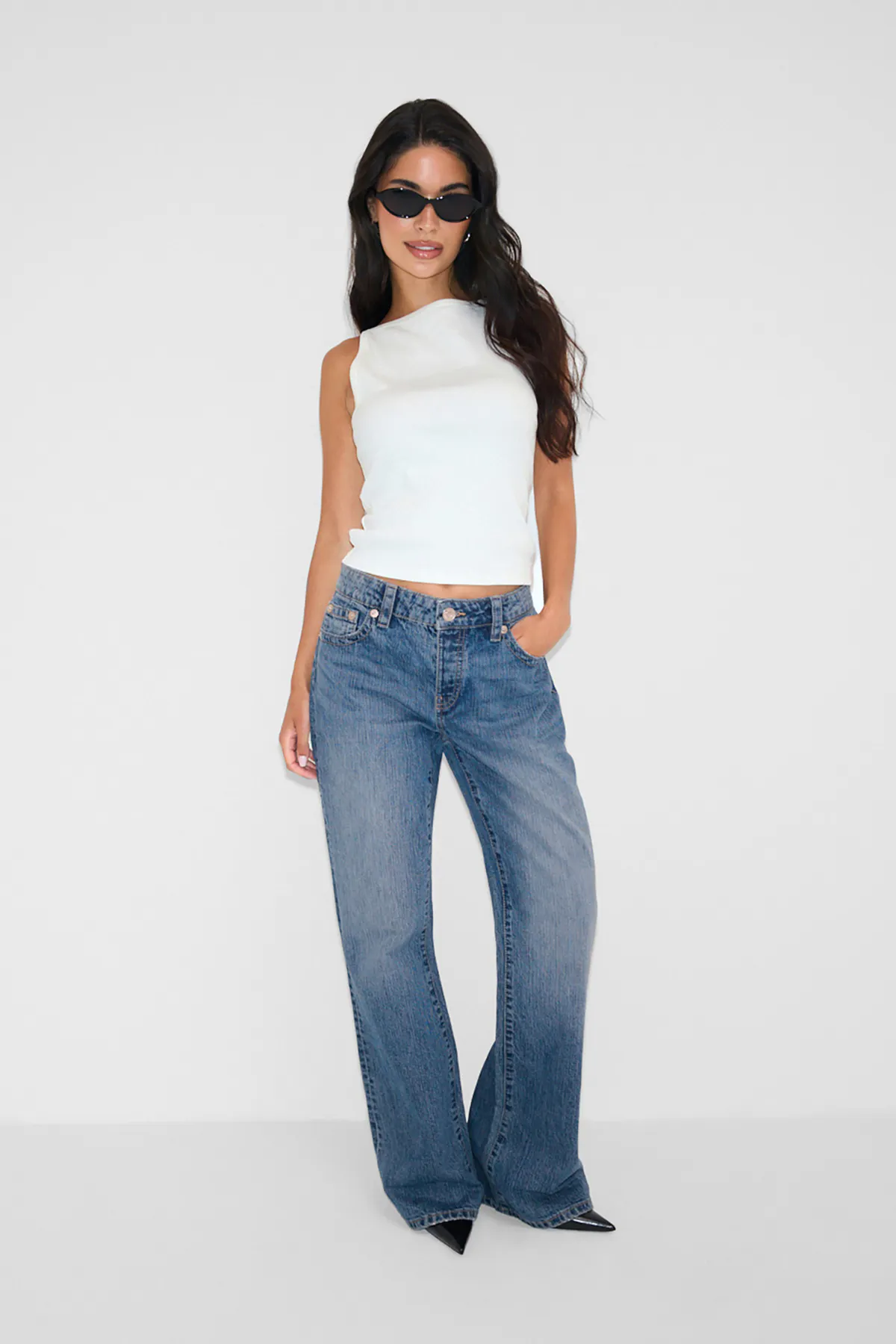Vit ärmlös topp med hög halsringning, avslappnade blå jeans med raka ben, svarta spetsiga klackar och svarta cat-eye-solglasögon.