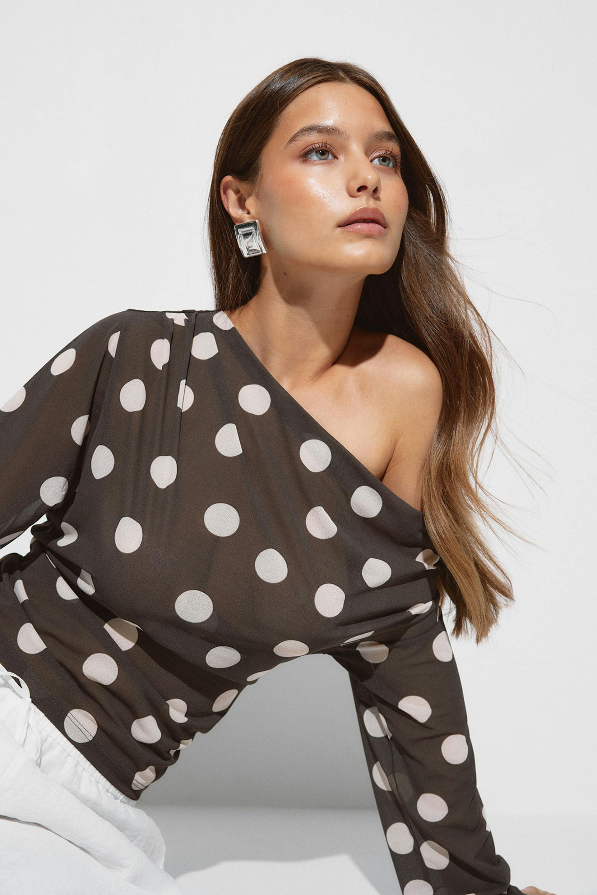 Blouse asymétrique à manches longues à pois foncés et gros points blancs, associée à un pantalon blanc et à des boucles d'oreilles carrées argentées surdimensionnées.