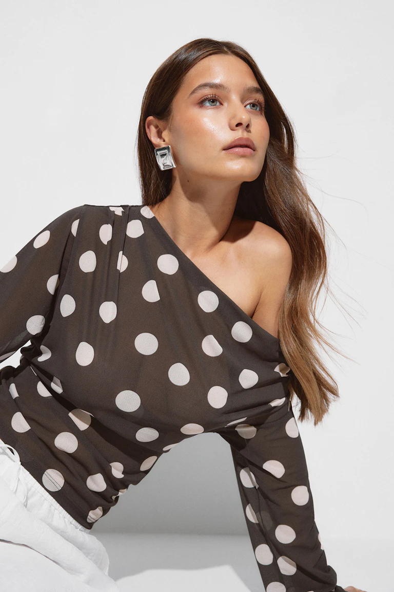 Asymmetrische, langärmelige, dunkle Polka-Dot-Bluse mit großen weißen Punkten, kombiniert mit einer weißen Hose und übergroßen, quadratischen Silberohrringen.
