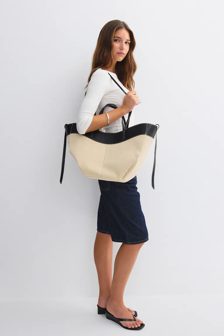 Modellen bär en vit, figurnära topp med trekvartsärm, knälång mörk jeanskjol, svarta lågklackade sandaler, en stor väska i beige canvas med svart läderkant samt ett silverfärgat armband.