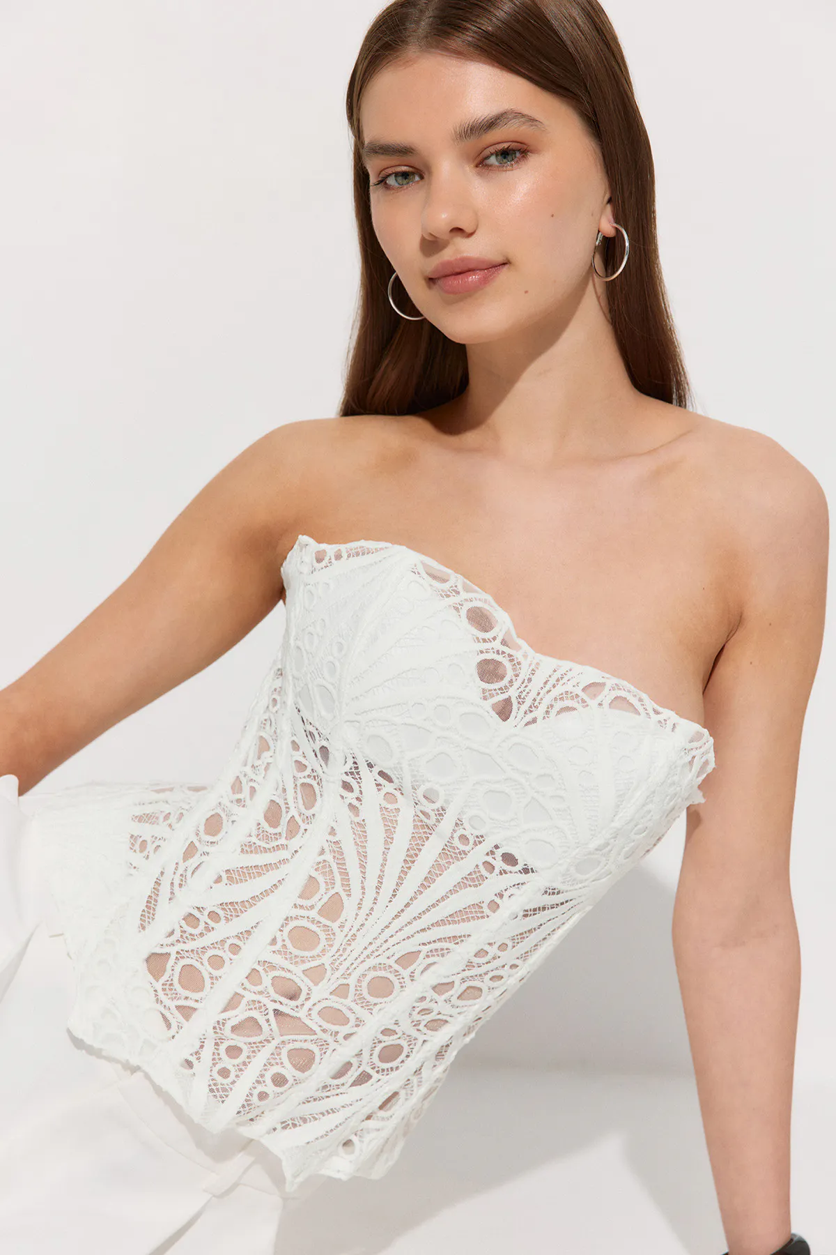 Strapless white lace corset with intricate cutout detailing, paired with white high-waist trousers and silver hoop earrings.