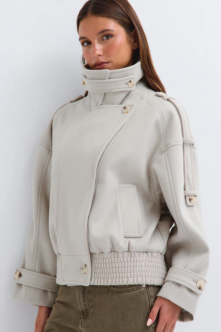 Hellbeige Jacke mit hohem Kragen und Knopfdetails, kombiniert mit einer olivgrünen Hose.