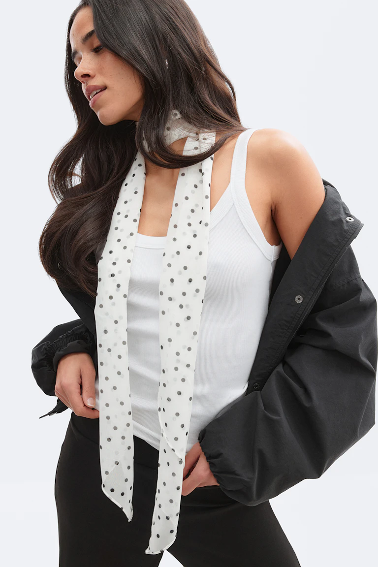 White ribbed tank top paired with a long white polka-dot scarf, a black oversized jacket worn off the shoulders, and black trousers.