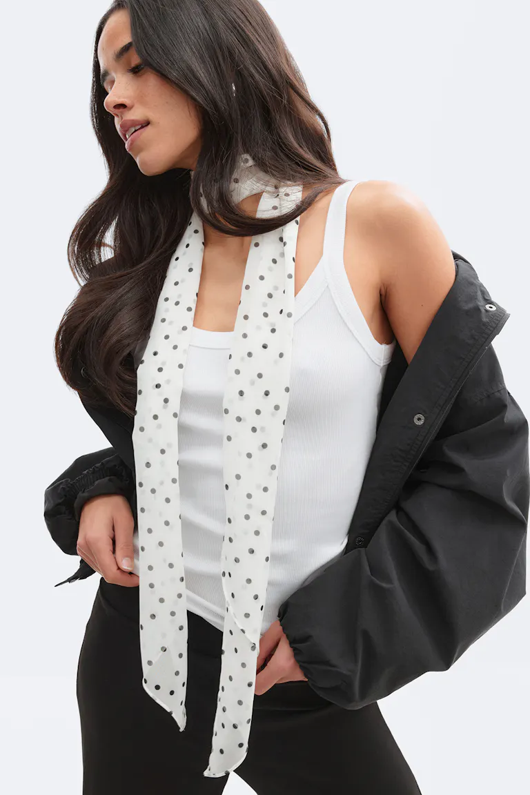 Hvit ribbestrikket tanktop kombinert med et langt, hvitt, prikkete skjerf, en svart, oversized jakke som bæres utenpå skuldrene, og svarte bukser.