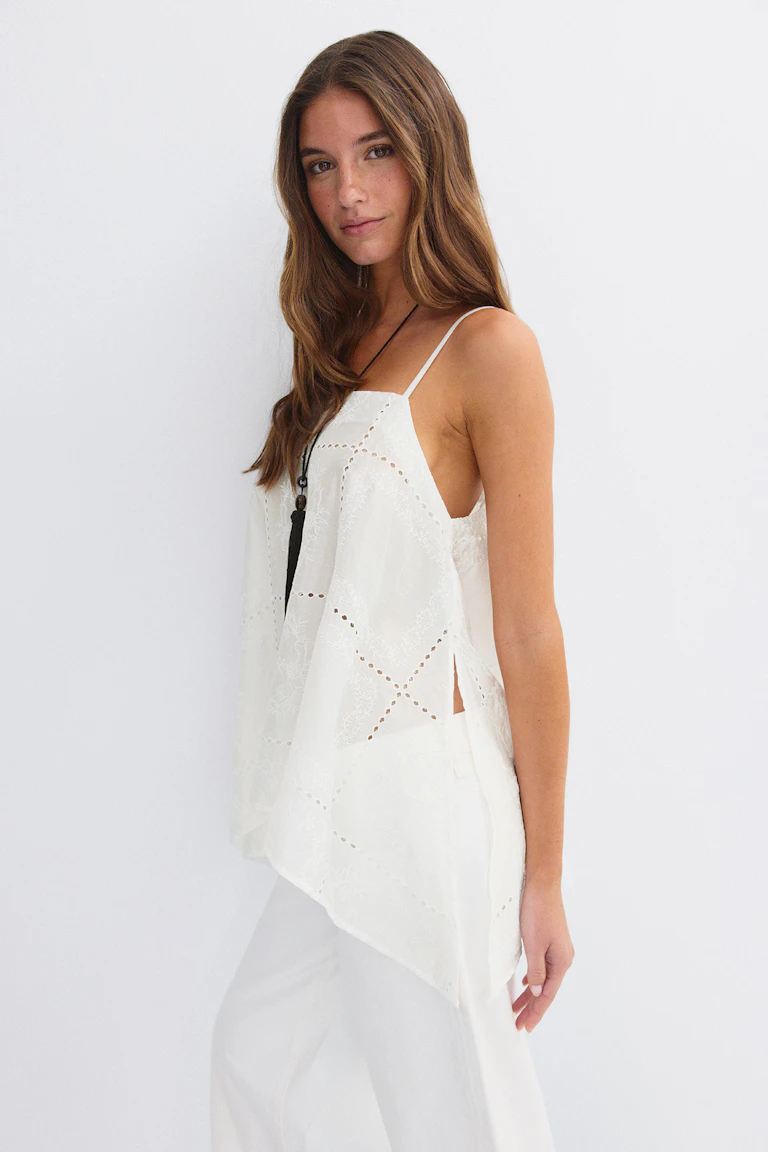 Caraco blanc en œillets brodés avec fines bretelles spaghetti, ourlet asymétrique et fente latérale, associé à un pantalon large blanc et à un long collier noir à pendentifs.